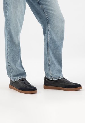 Person trägt hellblaue Jeans und schwarze Turnschuhe mit braunen Sohlen, steht auf einer weißen Fläche.