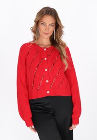 Piros kötött cropped kardigán ribbed mandzsettákkal, ferde fémes díszítéssel és nagy fehér gombokkal a záráshoz.