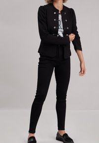 Veste noire sur mesure avec un col montant, des manches bouffantes et des boutons en métal argenté. Portée par-dessus un t-shirt à motif et associée à un jean noir.