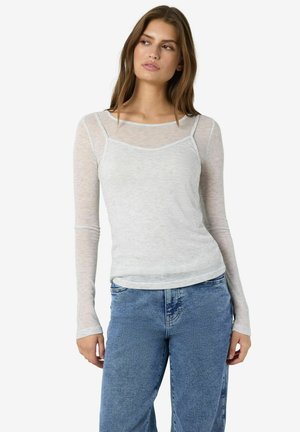Jonge vrouw in een doorschijnende witte top met lange mouwen, gelaagd met een witte hemdtop en een hoge taille spijkerbroek, kijkend opzij.