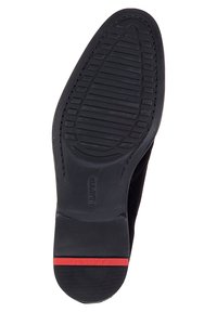Semelle en caoutchouc noir d'une chaussure présentant un motif de grip texturé, avec un logo central et une bande d'accent rouge au niveau du talon.