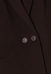 Veste croisée marron foncé au toucher doux, avec deux boutons ronds à l'avant et une poche inclinée sur le côté gauche.
