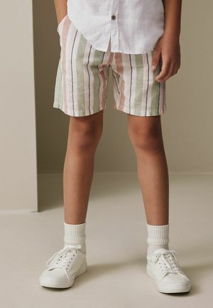 Barn iført pastelstribede shorts, hvide sokker, hvide sneakers og en delvist synlig hvid knaptrøje, stående på et beige gulv.