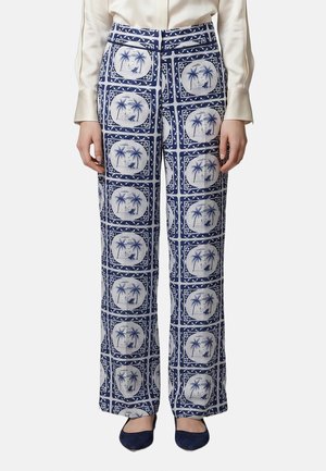 Vrouw draagt een broek met hoge taille en wijde pijpen met een blauw-witte palmboomprint, gecombineerd met een beige blouse en donkerblauwe platte schoenen.