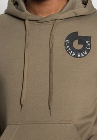 Sudadera caqui con cordón, bolsillo frontal y un logo circular negro con el texto "G.STAR RAW E.89" en el pecho. Material de suave mezcla de algodón.