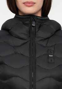 Black puffer jacket with a high collar, featuring a zip closure and a textured, wavy pattern. Logo tag on the left side near the zipper.