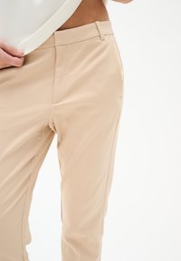 Béžové kalhoty na míru s hladkou texturou, bočními kapsami a úzkým pasem, s minimem švů pro elegantní vzhled.