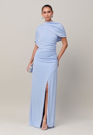 Vrouw draagt een lichtblauwe geplooide jurk met een asymmetrische split, houdt een kleine zilveren clutch vast en draagt zilveren sandalen met hoge hakken en oorbellen.