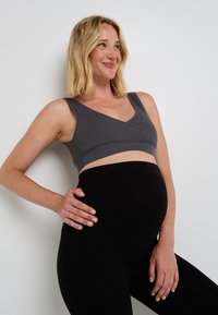 Soutien-gorge de sport de maternité gris à côtes avec décolleté en V et larges bretelles, associé à un legging noir taille haute, porté par une personne avec un ventre de femme enceinte visible.