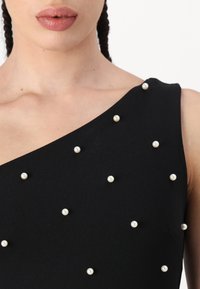Zwarte mouwloze jurk met een asymmetrische halslijn, versierd met kleine witte sierparels die gelijkmatig over de stof zijn verdeeld.