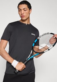 Melna sporta T-krekls ar "BOSS" logotipu; īsām piedurknēm, apaļu kakla izgriezumu. Modelis tur zilas un melnas krāsas tenisa riteni, valkā baltu aproci uz plaukstas.