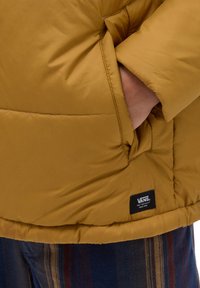 Chaqueta de plumas amarilla con una textura suave y brillante, que presenta un bolsillo lateral y una pequeña etiqueta de logo negra en la costura inferior.