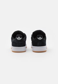 Chaussures de sport en daim noir avec semelle en caoutchouc blanche et accents, présentant une semelle extérieure en caoutchouc et un logo sur le talon.