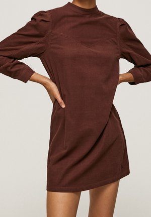 Nainen pukeutuneena ruskeaan, pitkähihaiseen polvipituiseen mekkoon, jossa on puffihihat, seisoo käsivarret lanteilla yksiväristä taustaa vasten.