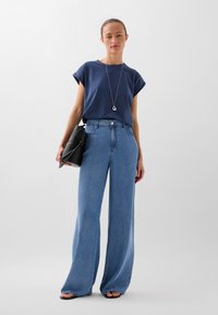Top navy a maniche corte con maniche arrotolate, abbinato a jeans denim a gamba larga di colore azzurro chiaro. Il modello tiene una borsa nera e indossa una collana d'argento.