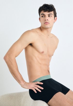 Čierne boxerky s mäkkým zeleným pásom, vyrobené z hladkého materiálu, ktorý sa prispôsobuje telu. Model sedí a ukazuje bočný profil.