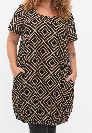 Personne aux cheveux bouclés portant une robe noire avec des motifs géométriques en diamants beiges, les mains dans les poches, et un tatouage floral visible sur l'avant-bras gauche.