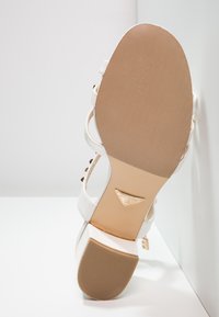 Valkoinen sandaali, jossa on useita hihnoja, teksturoitu beige pohja, kultaiset yksityiskohdat ja neliömäinen korko. Ominaisuuksiin kuuluu sileä päällinen ja koristeelliset niitit.