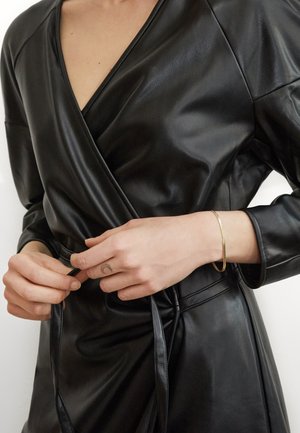 Schwarzes Leder-Wickelkleid mit V-Ausschnitt, langen Ärmeln und gebundenem Taillengürtel. Glatte Textur und minimalistisches Design, ergänzt durch ein goldenes Armband.