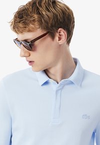 Nuori henkilö, jolla on lyhyet aaltoilevat hiukset, käyttää kilpikonnakehyksisiä aurinkolaseja ja vaaleansinistä polo-paitaa, katsoo alas ja sivuun.