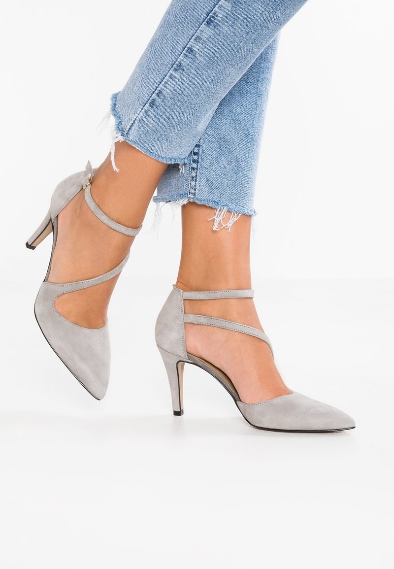Chaussures à talons hauts en daim gris avec brides à la cheville, portées avec un jean bleu clair à ourlet effiloché, jambes croisées sur fond blanc.