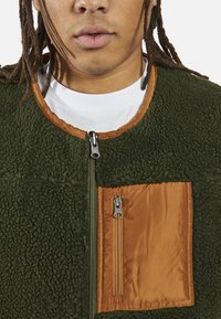 Casaco de felpa verde com um bolso de peito em satin laranja e fecho de correr. Gola arredondada com fecho de correr completo. Tecido texturizado.