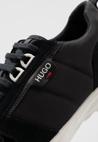 Närbild av svarta HUGO-sneakers med tyg- och mockatexturer, svarta snören, vita lufthål och en vit sula.