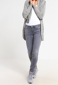 Grauer Cardigan mit offenem Vorderteil, über einem weißen Oberteil getragen. Slim-Fit graue Jeans und graue Sneaker runden das Outfit ab. Lässiger Stil.