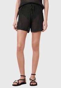 Zwarte gebreide shorts met een gestructureerd, open geweven patroon, voorzien van een trekkoord in de taille en zijzakken, gecombineerd met platte zwarte sandalen.