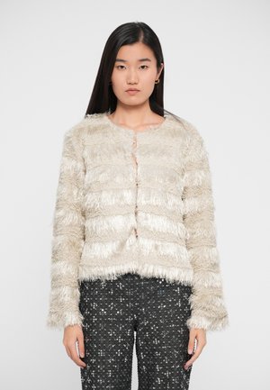Femme avec de longs cheveux noirs portant une veste frangée beige texturée et un pantalon noir à sequins, se tenant devant un fond blanc uni.