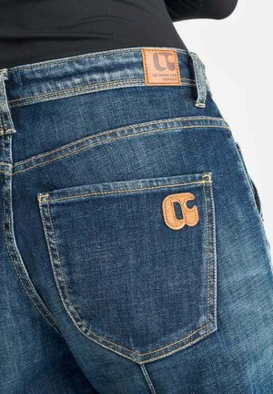 Gros plan sur une personne portant un jean en denim bleu avec des coutures visibles et des patchs en cuir brun de marque sur la taille et la poche arrière.