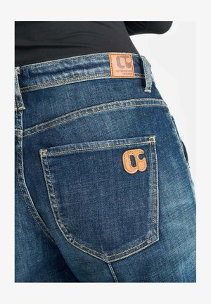 Primer plano de una persona que lleva vaqueros de mezclilla azul con costuras visibles y parches de cuero marrón de la marca en la cinturilla y en el bolsillo trasero.