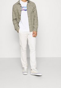 Ruudullinen vihreä ja musta flanellipaita valkoisen graafisen T-paidan päällä, yhdistettynä vaaleisiin chinopantsiin ja harmaisiin, beigelle värjättäihin lenkkareihin.