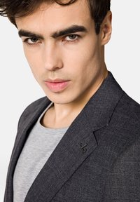 Ciemnoszary żakiet z teksturowanej tkaniny, z zapięciem na jeden guzik i małym czarnym logo na klapie, noszony na szarym koszuli.