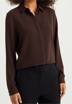 Personne portant une chemise marron foncé à manches longues et boutonnée, rentrée dans un pantalon noir taille haute, vue du cou aux hanches.