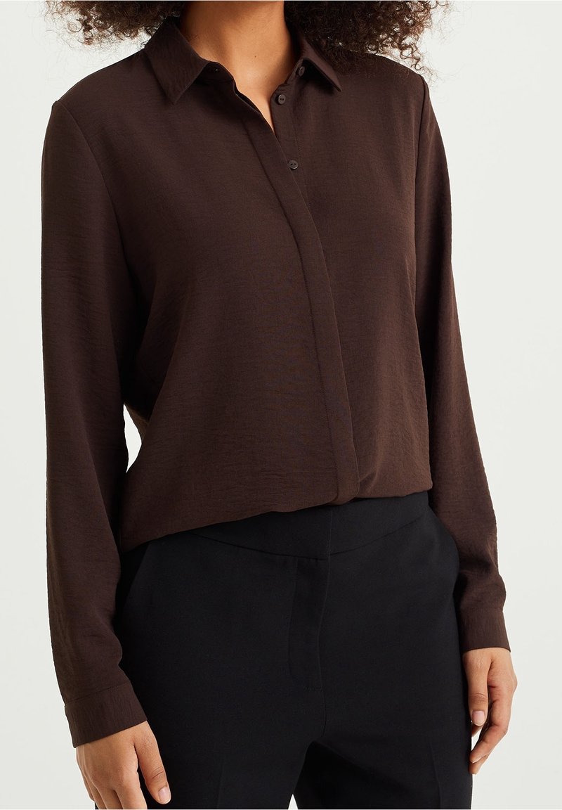 Personne portant une chemise marron foncé à manches longues et boutonnée, rentrée dans un pantalon noir taille haute, vue du cou aux hanches.