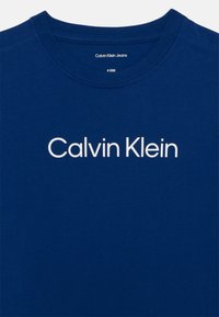 Plava pamučna majica s okruglim izrezom. Ima bijeli natpis "Calvin Klein" centriran sprijeda. Glatka tekstura, minimalistički dizajn.