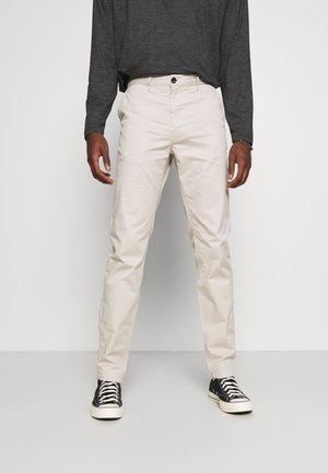 Chino in cotone beige con vestibilità dritta, dotati di chiusura a bottoni e tasche laterali, abbinati a scarpe da ginnastica nere e una camicia a maniche lunghe scura.