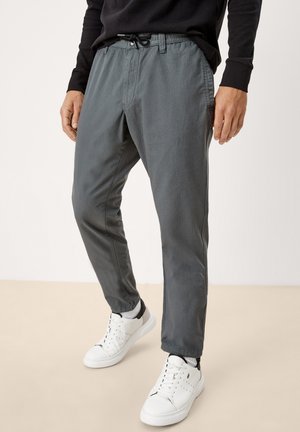 Homme portant un pantalon gris foncé à cordon, des baskets blanches avec des accents noirs et un haut noir à manches longues, debout sur un sol beige.