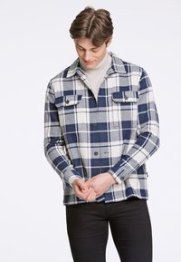 Camicia a quadri blu navy e crema con bottoni frontali, due tasche sul petto e colletto. Il tessuto appare texture e caldo.