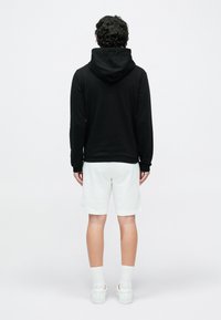 Osoba stojaca chrbtom, oblečená v čiernej mikine s kapucňou, bielych šortkách, bielych ponožkách a bielych teniskách na svetlom jednoduchom pozadí.