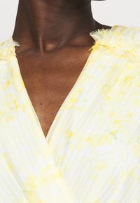 Ljust gult, veckat tyg med ett blommönster och fransar vid halsringningen. Designen har en V-ringning.