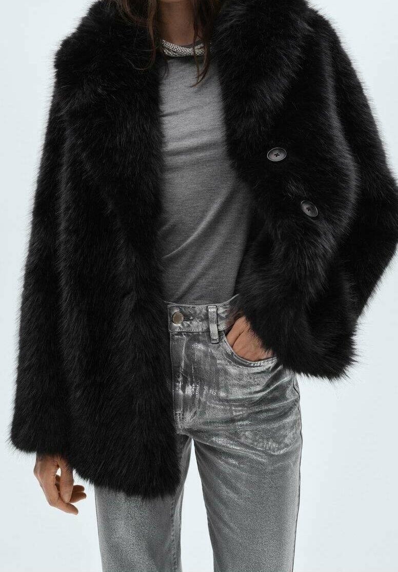 Femme portant une veste en fausse fourrure noire, un t-shirt gris, un jean métallique argenté et un collier chaîne argenté, main dans la poche.