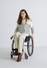 Hvit stripete genser med høy hals, beige vide bukser, brune ankelstøvler og en rullestol mot en ensfarget bakgrunn.