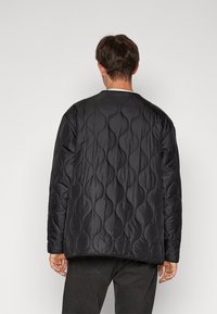 Black quilted jacket featuring a wavy pattern, round neckline, and relaxed fit, made from smooth, lightweight material with a matte finish.