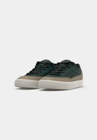 Green and beige sneakers with a suede and leather upper, flat laces, a padded collar, and a rubber outsole. Red logo accent on the side.