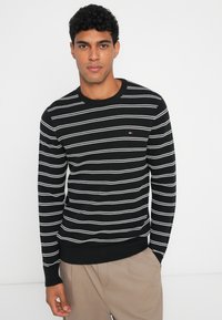Maglione nero con strisce orizzontali bianche, scollatura rotonda, polsini e orlo a costine. Piccolo logo sul petto, tessuto a maglia testurizzato.