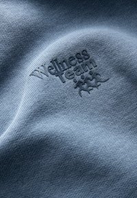 Jasnoniebieska tkanina o teksturowanym splocie z uniesionym, tonalnym logo "Zespół Wellness" oraz trzema stylizowanymi postaciami.