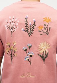 Ροζ ύφασμα με έξι πολύχρωμα κεντητά άγρια λουλούδια τοποθετημένα σε δύο σειρές και χειρόγραφο κέντημα "Les Deux" από κάτω.