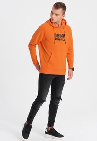 Felpa arancione con tasca frontale, cordini e testo nero. Abbinata a jeans skinny neri strappati e sneakers nere.